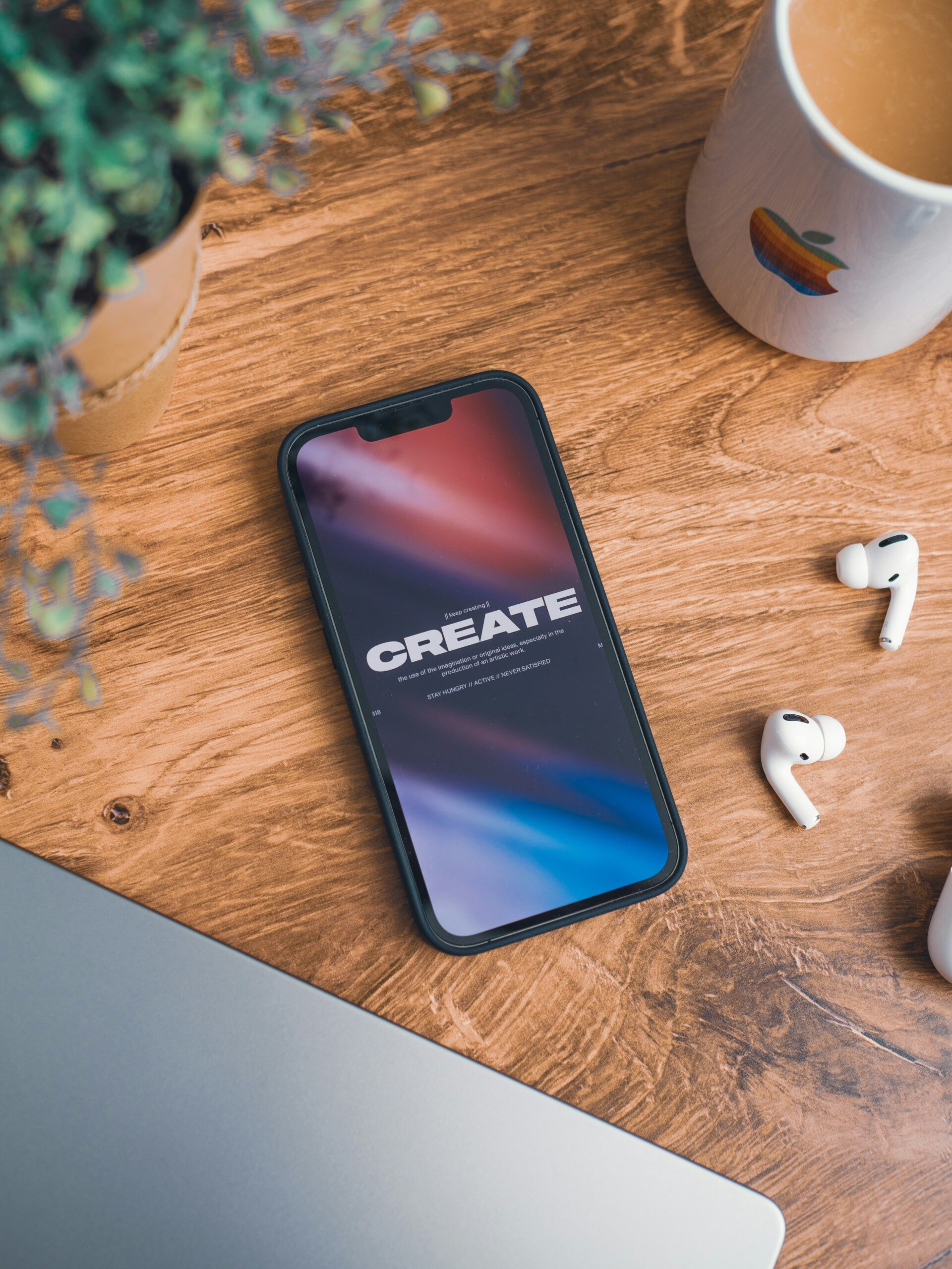 A phone lying face up on a desk, displaying the word 'create' on the screen, surrounded by headphones, a coffee mug, a plant, and part of a computer.